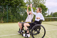 Person in wheelchair and standing friend celebrating success on tennis court